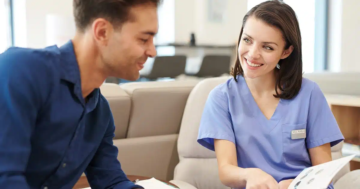 A healthcare professional wearing blue scrubs is smiling and discussing something with a patient who is sitting across from her.