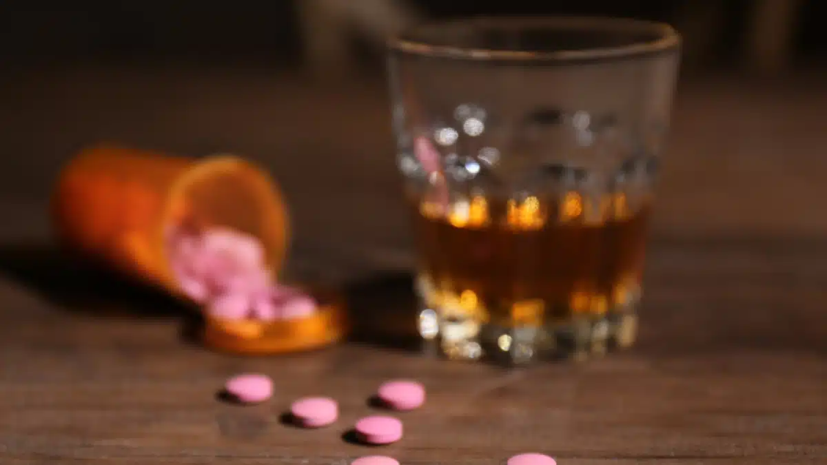 Pills spilled on a table next to an alcoholic beverage. Graphic explains the safe time to wait before drinking depends on your prescription