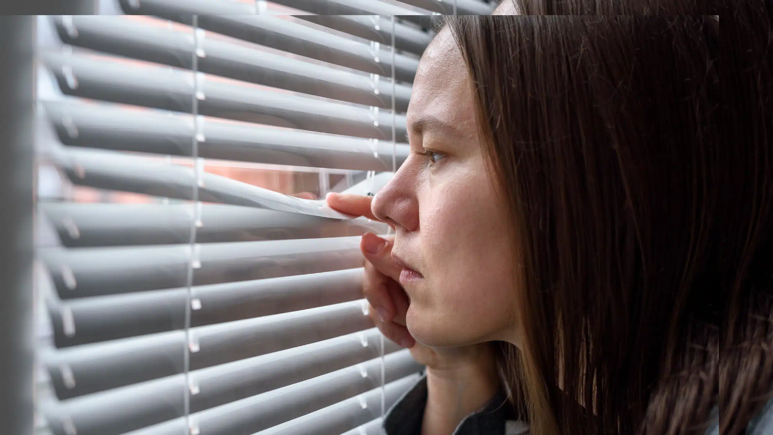 Woman peering out her window through the blinds. Text explains how anxiety can produce physical effects