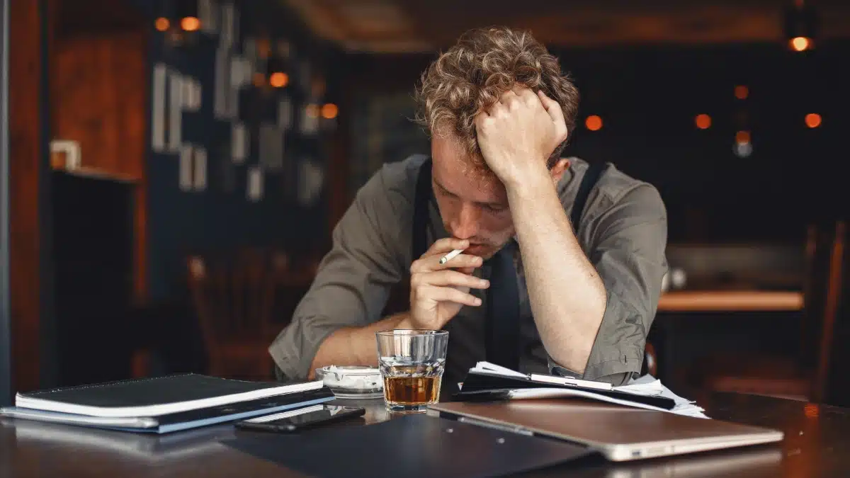 Man smoking a cigarette with a glass of whisky in front of him with his head resting in his hand. Both anxiety and addiction can coexist.