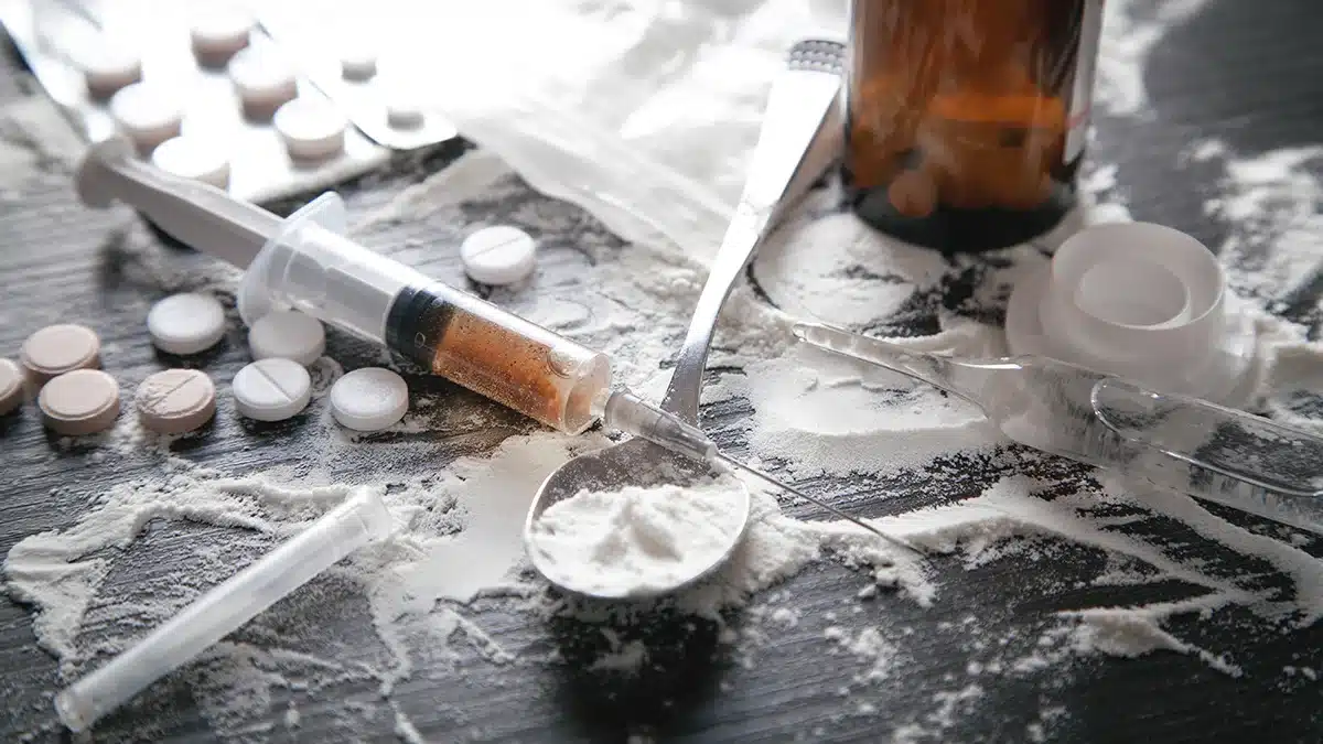 drug paraphernalia including a syringe, spoon with white powder, pills, and glass vials scattered across a dark surface.